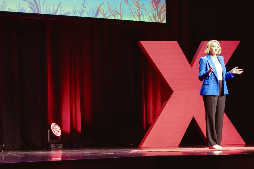 Jo Kelly stands on the TEDxBrisbane stage in a blue jacket and black trousers